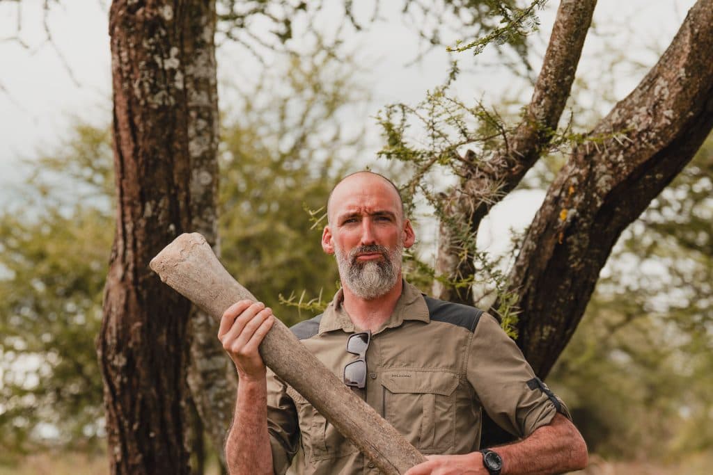 Een man met een kaal hoofd en grijze baard staat buiten tussen de bomen in Afrika en houdt een groot dierenbot vast. Hij draagt een kaki overhemd, een zonnebril hangt aan zijn kraag en hij heeft een ernstige uitdrukking.
