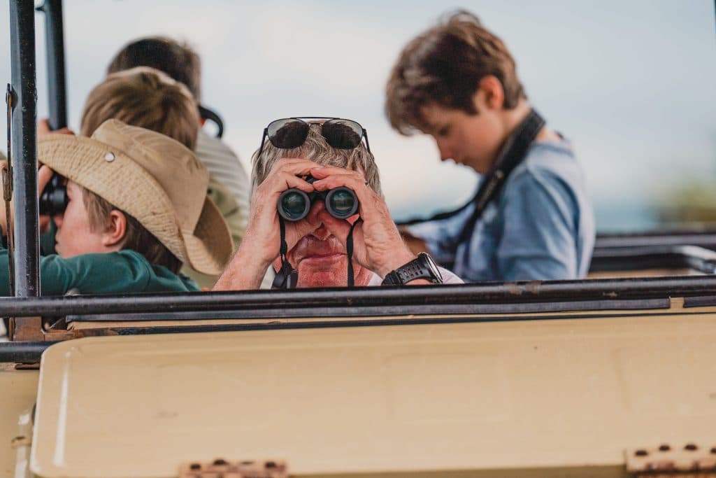 Een groep mensen in een safarivoertuig in Tanzania, met één persoon in het midden die door een verrekijker kijkt, anderen met hoeden op die in verschillende richtingen kijken, genietend van het paaltjesritme van een authentiek Afrika-avontuur.