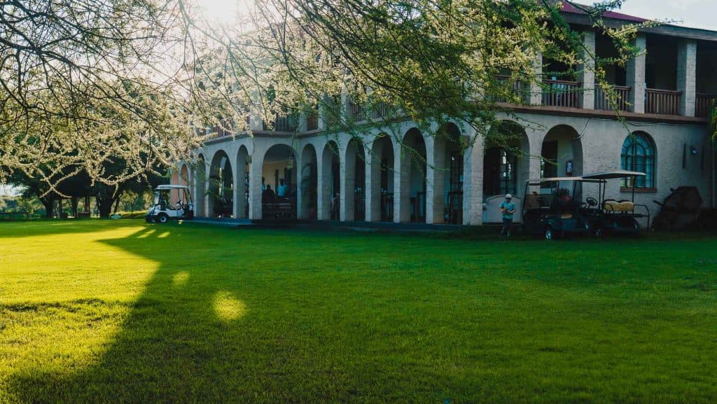 Zonlicht filtert door boomtakken over een groot gebouw met gewelfde zuilen, een balkon en golfkarretjes geparkeerd op het groene gazon aan de voorkant. Dit biedt een rustige glimp van Afrika, waar het leven zich ontvouwt aan de deur van paal Tanzania.