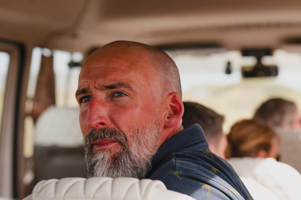 Een man met een kaalgeschoren hoofd en grijze baard zit in een voertuig en kijkt achterom over zijn schouder. Zonlicht belicht zijn gezicht terwijl hij door Tanzania hoogtepunten rijdt, met andere passagiers zichtbaar op de achtergrond.