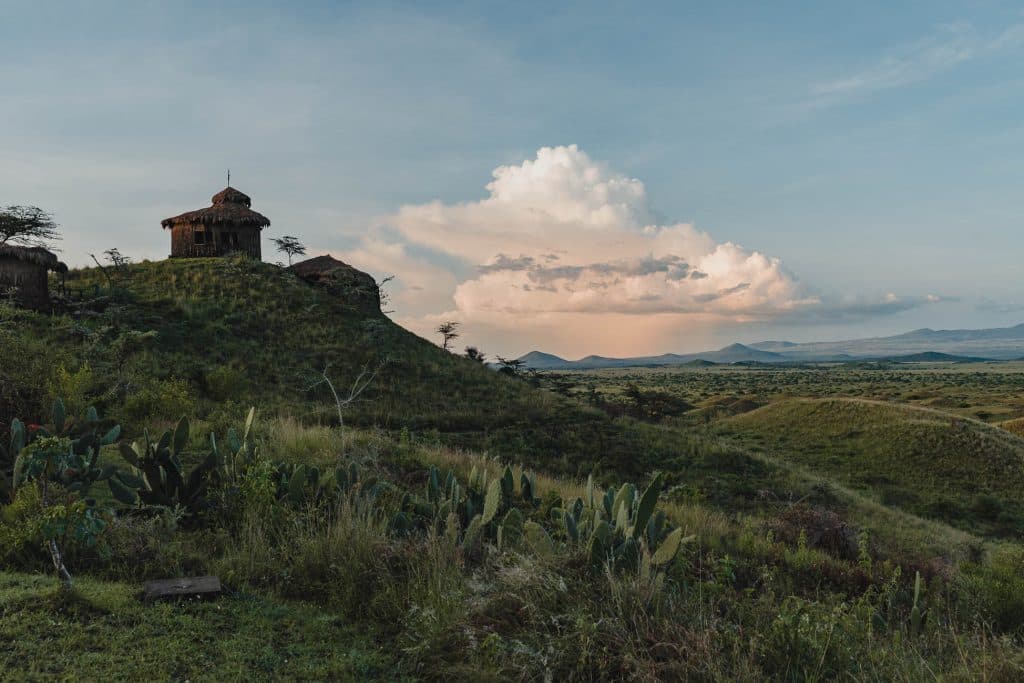 Een rustiek stenen gebouw staat op een groene heuvel onder een blauwe hemel met grote wolken, met uitzicht op een uitgestrekt, open savannelandschap - een van de echte hoogtepunten van Tanzania, met bergen in de verte en verspreide vegetatie die het adembenemende tafereel compleet maken.