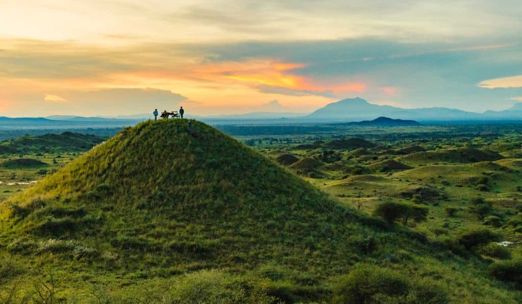 Een groep mensen staat op de top van een met gras begroeide heuvel bij zonsondergang, uitkijkend over een uitgestrekt landschap met glooiende heuvels en bergen in de verte - een van de adembenemende hoogtepunten van Tanzania onder een kleurrijke hemel.