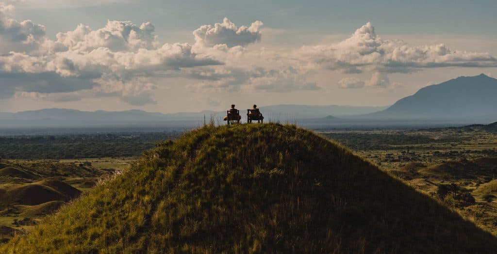 Twee mensen zitten op bankjes bovenop een met gras begroeide heuvel en kijken uit over een uitgestrekt landschap met bergen in de verte - een tafereel dat doet denken aan hoogtepunten in Tanzania - onder een dramatische, wolkenlucht.