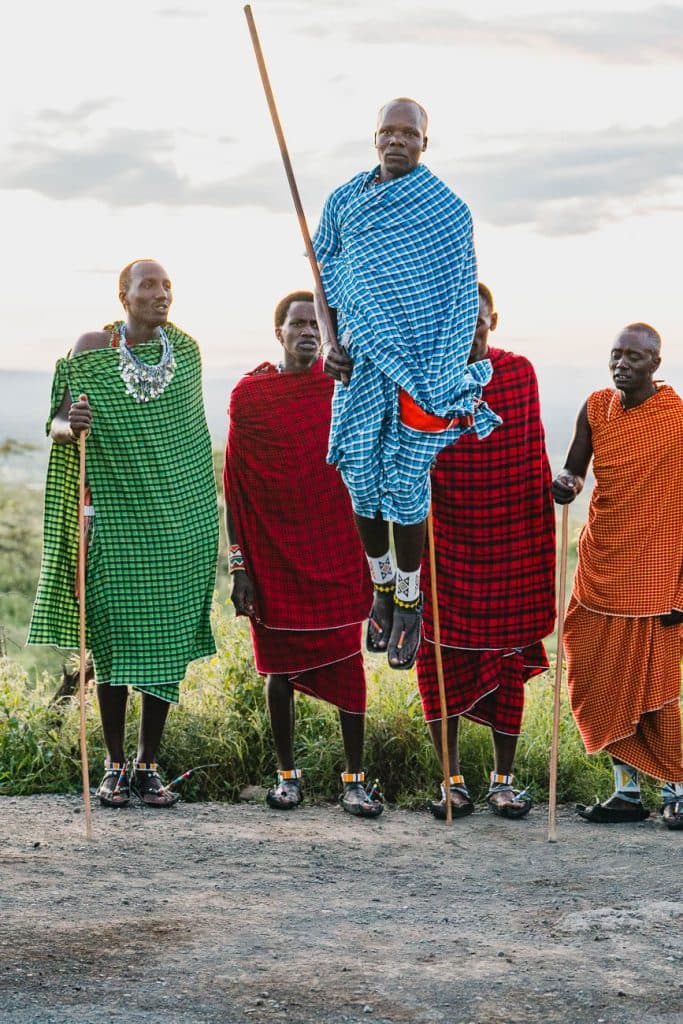 Vijf Maasai-mannen in kleurrijke traditionele shuka's staan buiten. Eén man springt met een staf in de lucht terwijl de anderen toekijken. Afgezet tegen weelderig groen en een heldere hemel, is deze scène een beeld van een van de authentieke hoogtepunten van Tanzania.