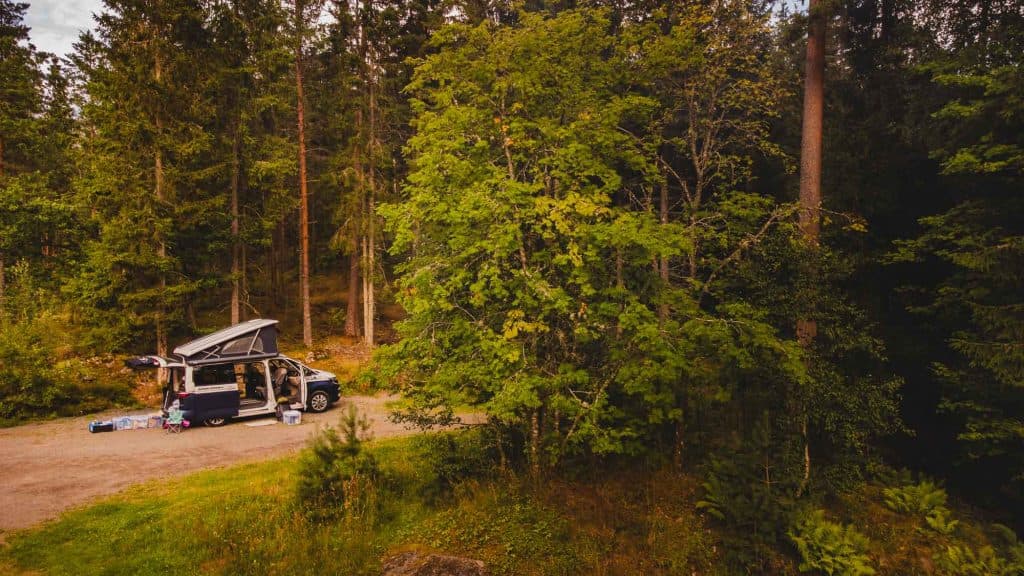 Een camper met het dak omhoog staat geparkeerd op een onverharde weg in een dicht bos in Zweden, met buiten kampeerspullen klaargezet. Weelderige groene bomen omringen de vredige, openluchtscène - perfect om het naturisme te midden van de natuur te omarmen.
