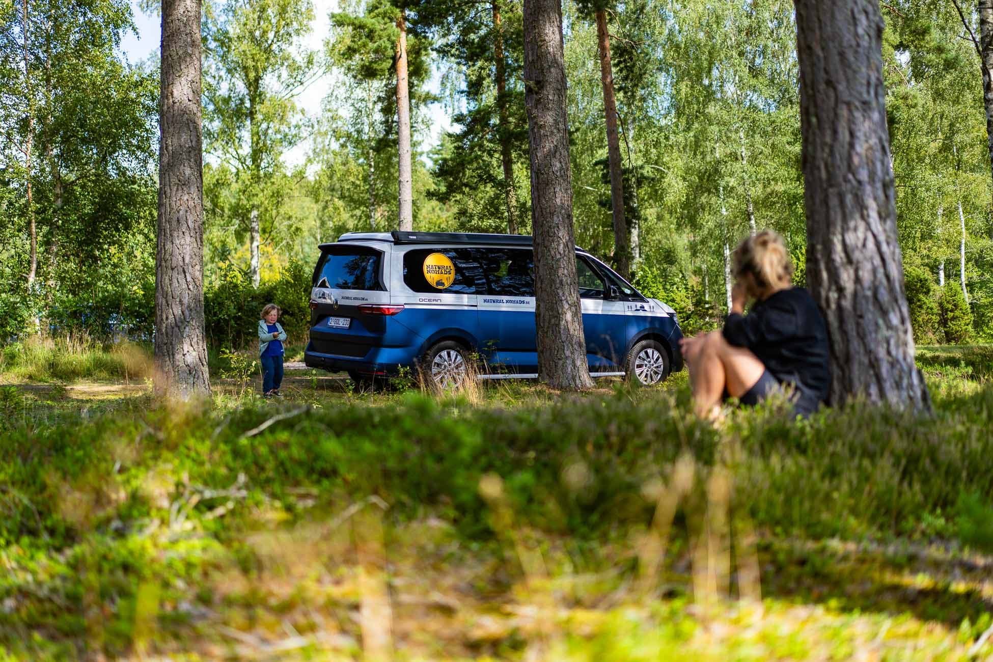 Een blauwe camper staat geparkeerd tussen hoge bomen in een Zweeds bos. Een kind staat bij het busje terwijl een volwassene op het gras op de voorgrond zit en naar het kind kijkt. Zonlicht filtert door de bomen en creëert een serene naturistische sfeer.
