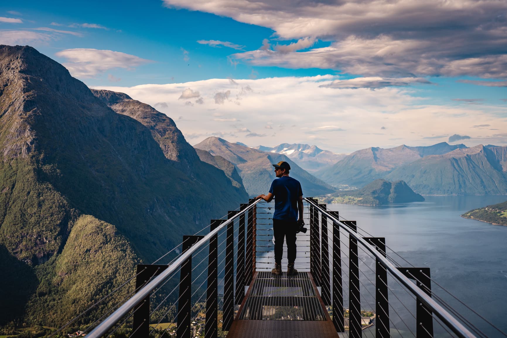 Iemand staat op een schilderachtig uitkijkplatform dat uitkijkt over de dramatische fjorden, omringd door hoge, groene bergen en water onder een gedeeltelijk bewolkte hemel - een perfect tafereel voor Noorwegen Nomads die de middernachtzon najagen.