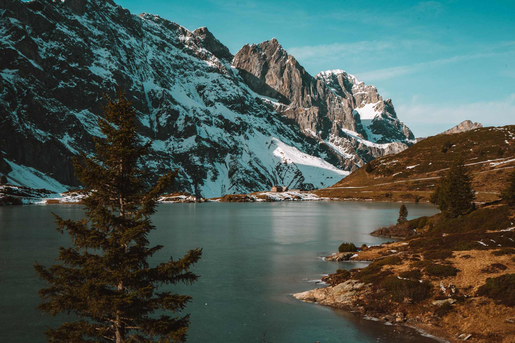 Een rustig meer weerspiegelt besneeuwde bergen onder een blauwe hemel, met altijd groene bomen en rotsachtig terrein op de voorgrond - perfect voor een herfst citytrip in Zwitserland.