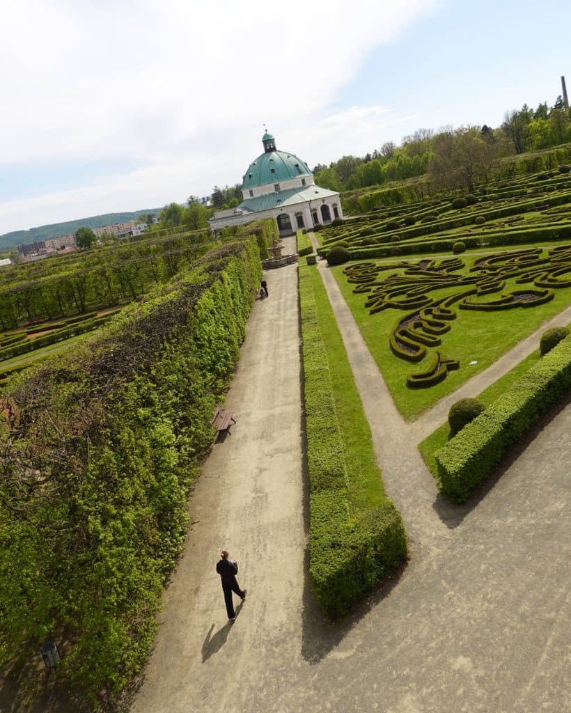 Een persoon loopt over een breed pad omzoomd door gemanicuurde heggen in een formele tuin. Sierlijke geometrische patronen versieren het gazon, terwijl een groot wit paviljoen dat doet denken aan een paleis in Tsjechië op de achtergrond staat onder een blauwe hemel.