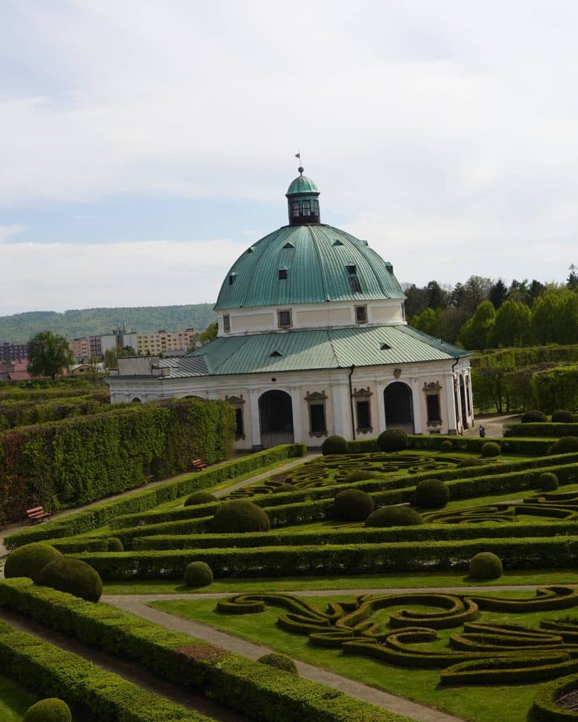 Een koepelvormig, wit barok paviljoen met een groen koperen dak staat te midden van formele vormsnoeituinen in Tsjechië, waar ingewikkelde hagen de elegantie van een prinses oproepen. Banken en hoge gesnoeide heggen omzomen de tuin; bomen en gebouwen in de verte maken het uitzicht compleet.