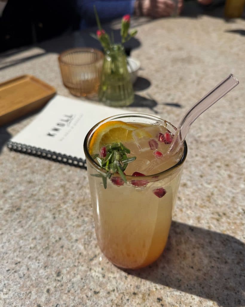Een glas ijskoude limonade met een glazen rietje, gegarneerd met citroen, rozemarijn en granaatappelpitjes, staat op een zonovergoten stenen tafel.