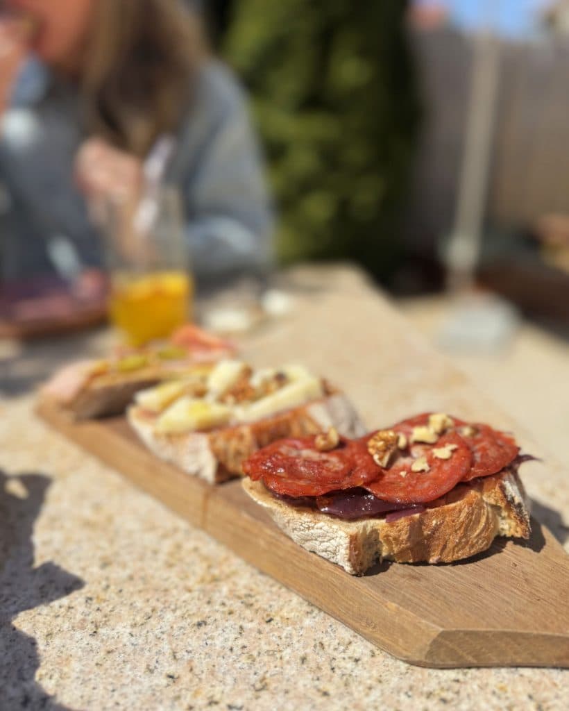 Drie sneetjes rustiek brood belegd met vleeswaren, kaas en walnoten rusten op een houten plank. Op de achtergrond geniet iemand van een voordelige lunch buiten in de zon, drinkend uit een glazen fles, terwijl hij zich misschien in Tsjechië waant.