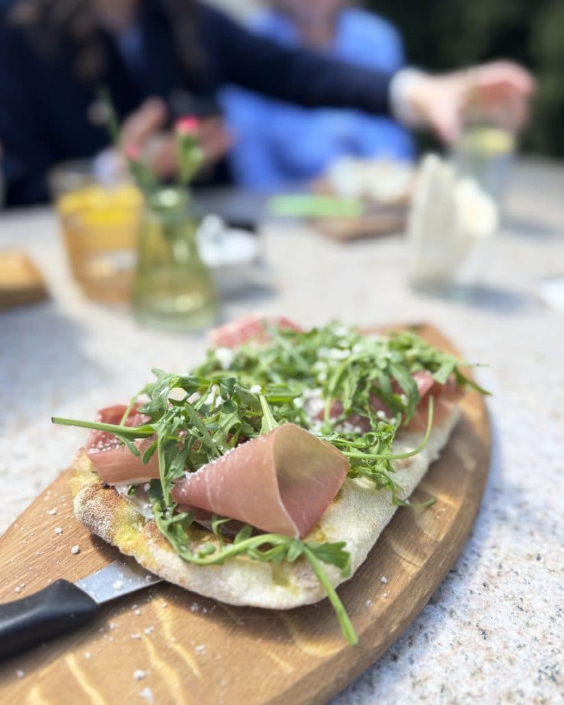 Een close-up van een houten plank met platbrood belegd met prosciutto, rucola en geraspte kaas. Op de onscherpe achtergrond zitten mensen aan een tafel in Tsjechië, drankjes in de hand, zonlicht schijnt op bloemen en er wordt vrolijk gelachen.