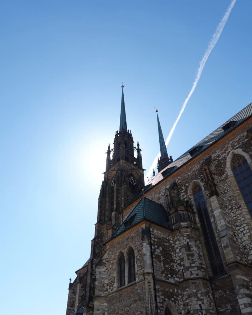 Hoge gotische kerk met twee spitse torenspitsen tegen een helderblauwe lucht in Brno, Tsjechië. De zon achter de centrale torenspits creëert een halo-effect, terwijl een witte contrail boven het gedetailleerde steenwerk en de boogramen kruist.