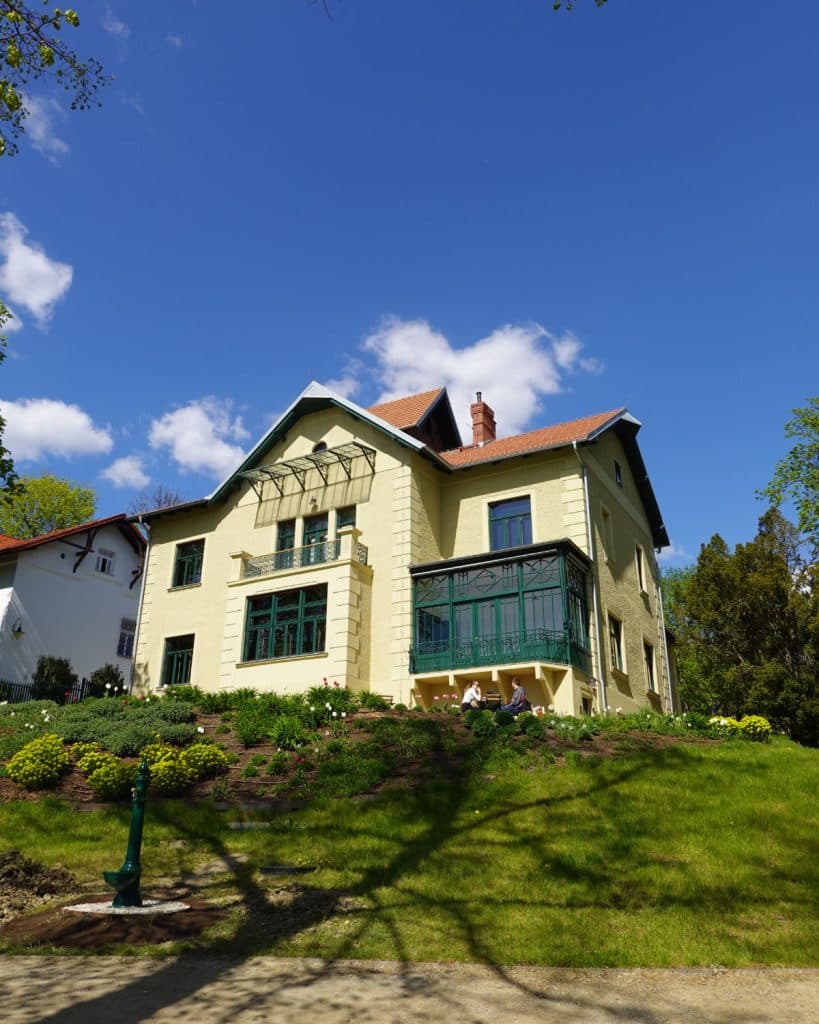Een geel huis van twee verdiepingen met groene raamkozijnen en een balkon staat op een met gras begroeide heuvel in Brno onder een helderblauwe lucht met verspreide wolken. Struiken, bloemen en een groene lantaarnpaal sieren de tuin, met links een ander wit huis.