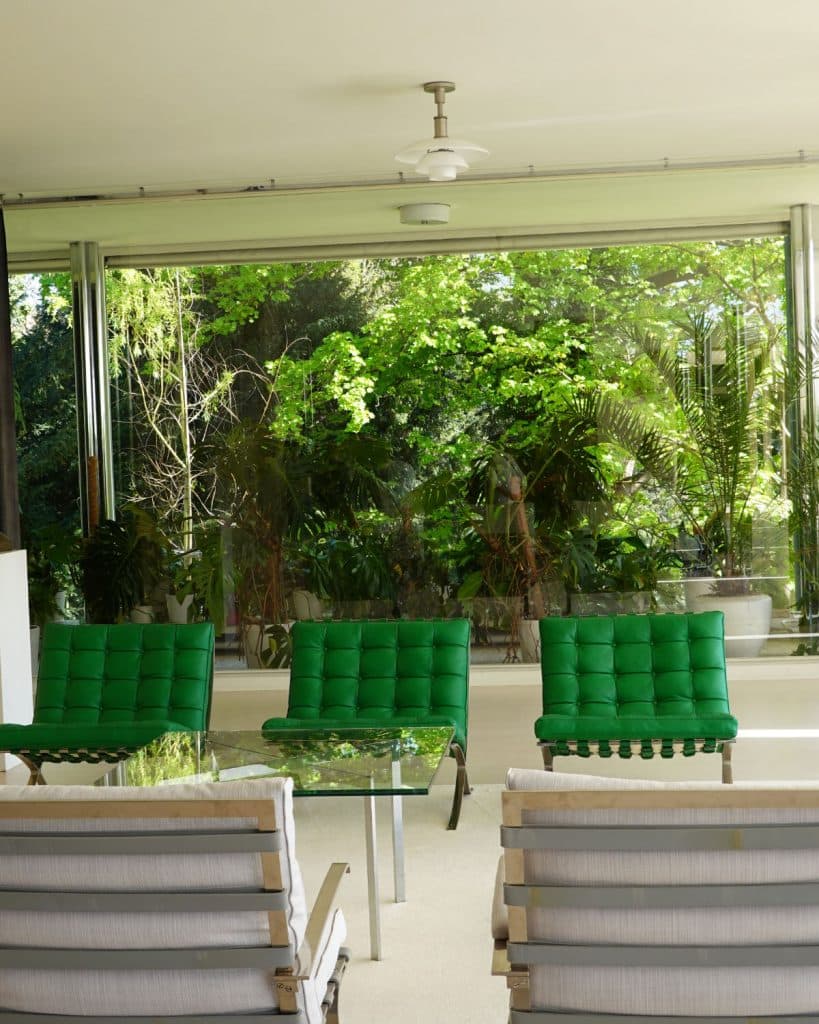 Moderne, minimalistische kamer met grote glazen ramen die de weelderige groene planten buiten laten zien. Binnen staan vier felgroene, getufte stoelen tegenover een glazen salontafel - een stijlvolle ruimte die perfect is voor reisinspiratie of om te dromen van Brno op het Tsjechische platteland.
