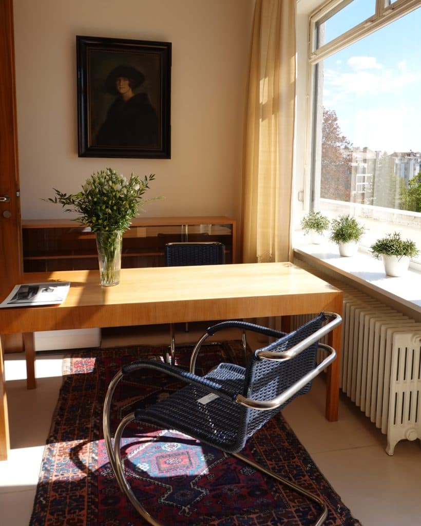 Een zonovergoten kantoor met een houten bureau, een vaas met groene takken en een navy geweven schommelstoel met metalen frame roept de charme op van de Tsjechische stijl in Brno. Een kleurrijk tapijt, potplanten op de vensterbank en een donker portret maken deze uitnodigende, op reizen geïnspireerde ruimte compleet.