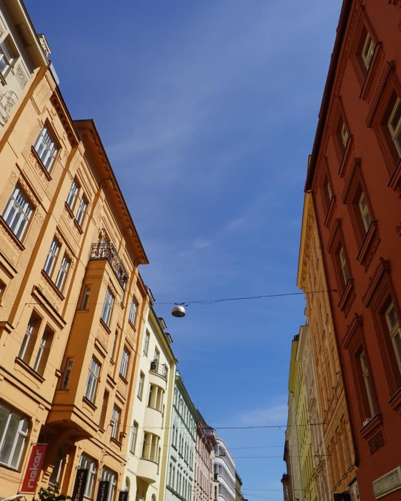 Hoge, kleurrijke gebouwen met sierlijke details staan aan weerszijden van een smalle straat in Brno, Tsjechië. De gevels - oranje, geel, groen en rood - betoveren reisliefhebbers onder een straatlantaarn en een helderblauwe lucht met pluizige wolken.