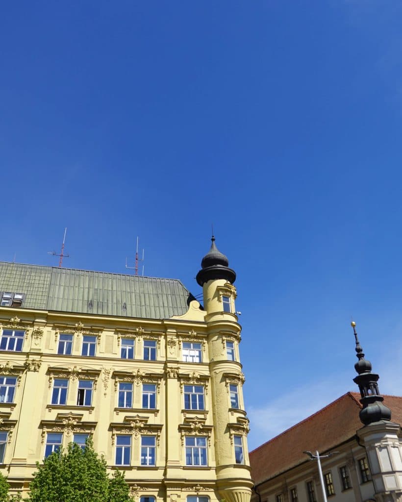 Een geel historisch gebouw met sierlijke details en een kleine zwarte koepel staat onder een helderblauwe hemel in Brno, Tsjechië. Een deel van een ander gebouw met een rood dak en donkere torenspits is zichtbaar aan de rechterkant, omlijst door groene boomtoppen - een reispareltje.