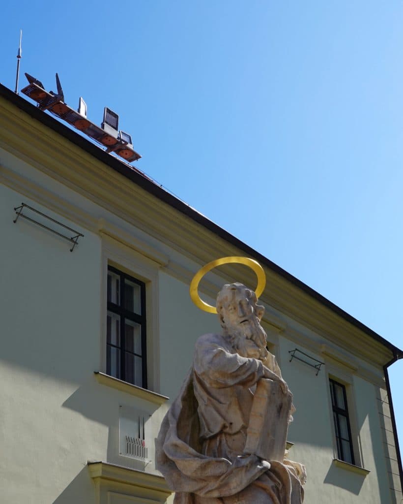 Een stenen standbeeld van een bebaarde man met een geel aureool staat voor een lichtgeel gebouw in Brno, Tsjechië. Op de voorgrond staat het standbeeld, terwijl zonnepanelen op het dak deze unieke reisbestemming onder een strakblauwe hemel accentueren.