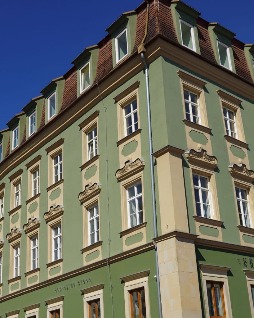 Een groen, drie verdiepingen tellend historisch gebouw in Brno, Tsjechië, met witomrande ramen, decoratieve beige accenten en een bruin, betegeld mansardedak steekt af tegen een helderblauwe hemel - een uitnodigend gezicht voor elke reisliefhebber.