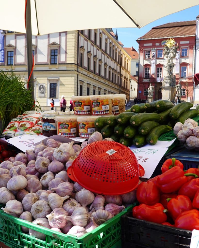 Een marktkraam in Brno toont knoflookbollen, rode paprika's, courgettes, potten jam en kaaswielen onder een witte paraplu. Gebouwen en een standbeeld dat typerend is voor Tsjechië zijn op deze zonnige dag vlakbij te zien.