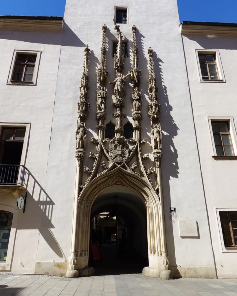 Een hoge, sierlijke gotische boog siert een bleek gebouw in Brno, Tsjechië. Ingewikkeld steenhouwwerk en een wapenschild bekronen de ingang, terwijl het zonlicht scherpe schaduwen werpt, perfect voor liefhebbers van reisfotografie.