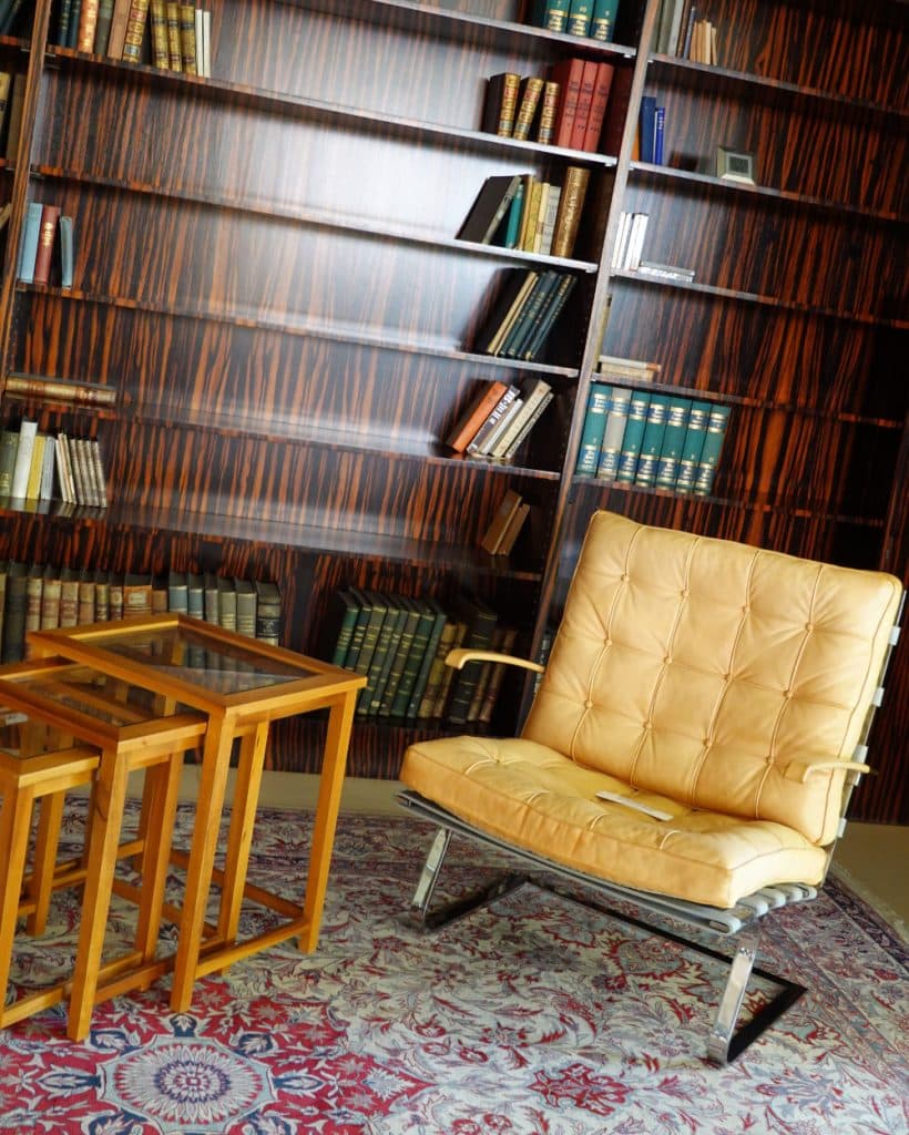 Een bruine leren loungestoel staat op een gedessineerd tapijt naast drie in elkaar geschoven houten tafels. Daarachter staan donkere, glanzende boekenplanken vol met boeken uit Brno en daarbuiten, waardoor deze gezellige studeerkamer een subtiele, op reizen geïnspireerde charme uitstraalt.