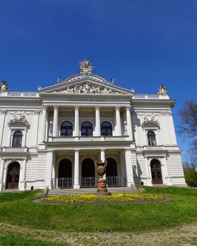 Een groots wit neoklassiek gebouw met zuilen, sierlijk houtsnijwerk en beelden op het dak staat in Brno, Tsjechië. Op de gevel staat "SLASKY DIVADLO". Een ronde tuin met gele bloemen en een decoratieve urn ligt ervoor onder een helderblauwe hemel.