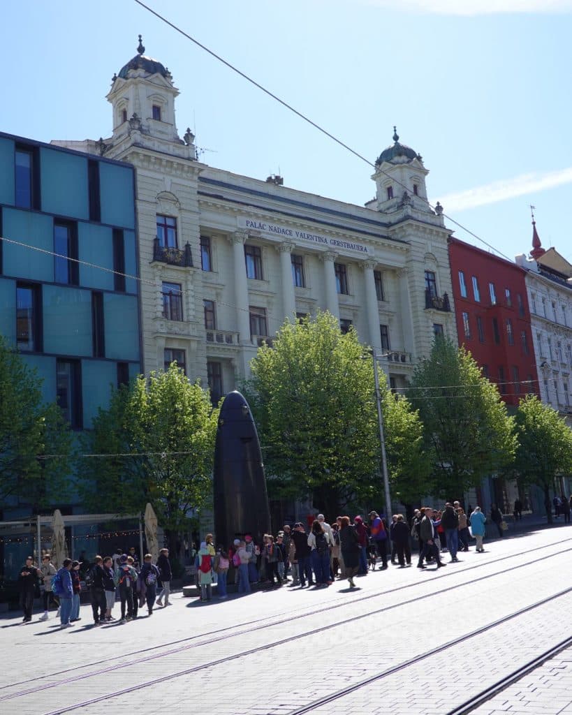 Een groep mensen staat in de rij bij een hoog, zwart, kogelvormig beeld op een geplaveide straat in Brno, Tsjechië. Op de achtergrond staan moderne glazen gebouwen, sierlijke architectuur met zuilen en bomen met felgroene bladeren.