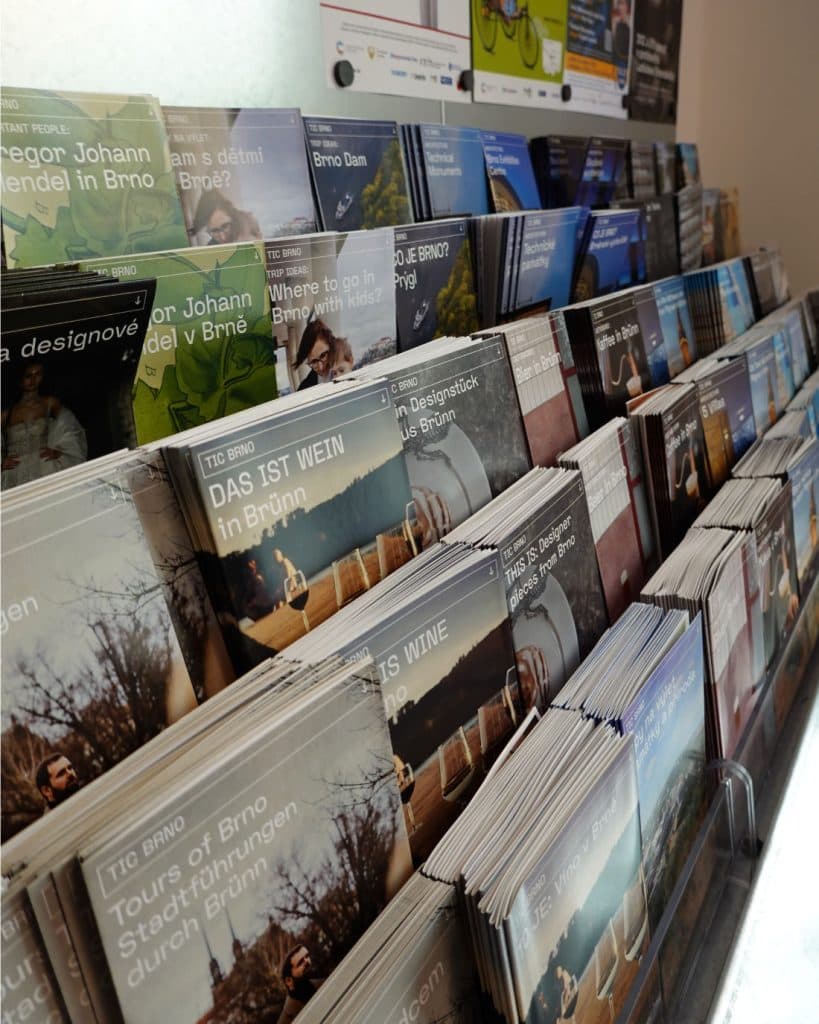 Een displayrek vol reisbrochures en pamfletten in meerdere rijen toont Tsjechische toeristengidsen voor Brno, met natuurfoto's, hoogtepunten van de stad en tweetalige titels. Het rek staat op een aanrecht in een goed verlichte omgeving.