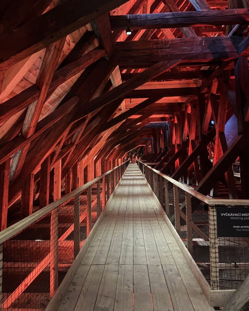 Een lange houten loopbrug met metalen leuningen strekt zich in de verte uit binnen een structuur van donkere houten balken in Brno, Tsjechië, alles verlicht door dramatische rode verlichting die schaduwen werpt en het geometrische raamwerk erboven benadrukt - een architectonische droom voor reizigers.