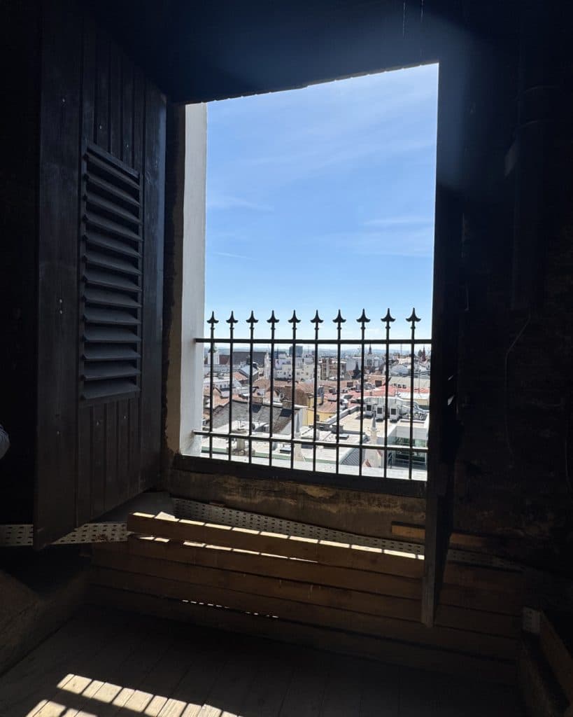 Een helder stadslandschap in Brno is zichtbaar door een hoog raam met metalen tralies. Het raamkozijn en de houten luiken liggen in de schaduw en contrasteren met de zonovergoten rode daken en de blauwe Tsjechische lucht. Onder het raam staan houten banken die uitnodigen tot dagdromen over reizen.