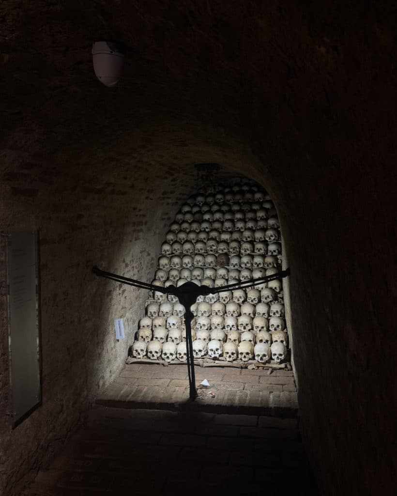 Een zwak verlichte, smalle stenen gang in Brno, Tsjechië toont een muur vol menselijke schedels achter een zwarte metalen barrière. De schedels liggen in rijen en vullen een nis. Schaduwen verdiepen de sfeer van deze ossuariumscène.