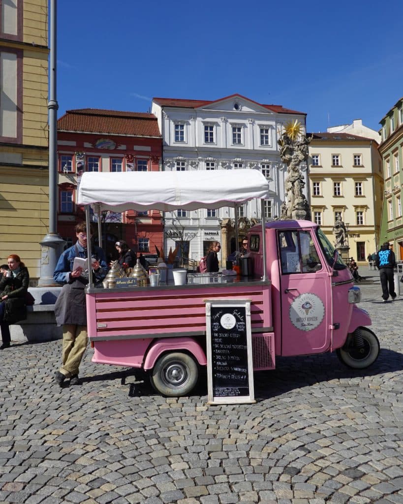 Een roze ijskar met een witte luifel staat geparkeerd op een geplaveid plein in Brno. Een man staat achter de toonbank terwijl mensen langslopen, omringd door kleurrijke oude Tsjechische gebouwen, een stenen standbeeld en een krijtbord met een menu.