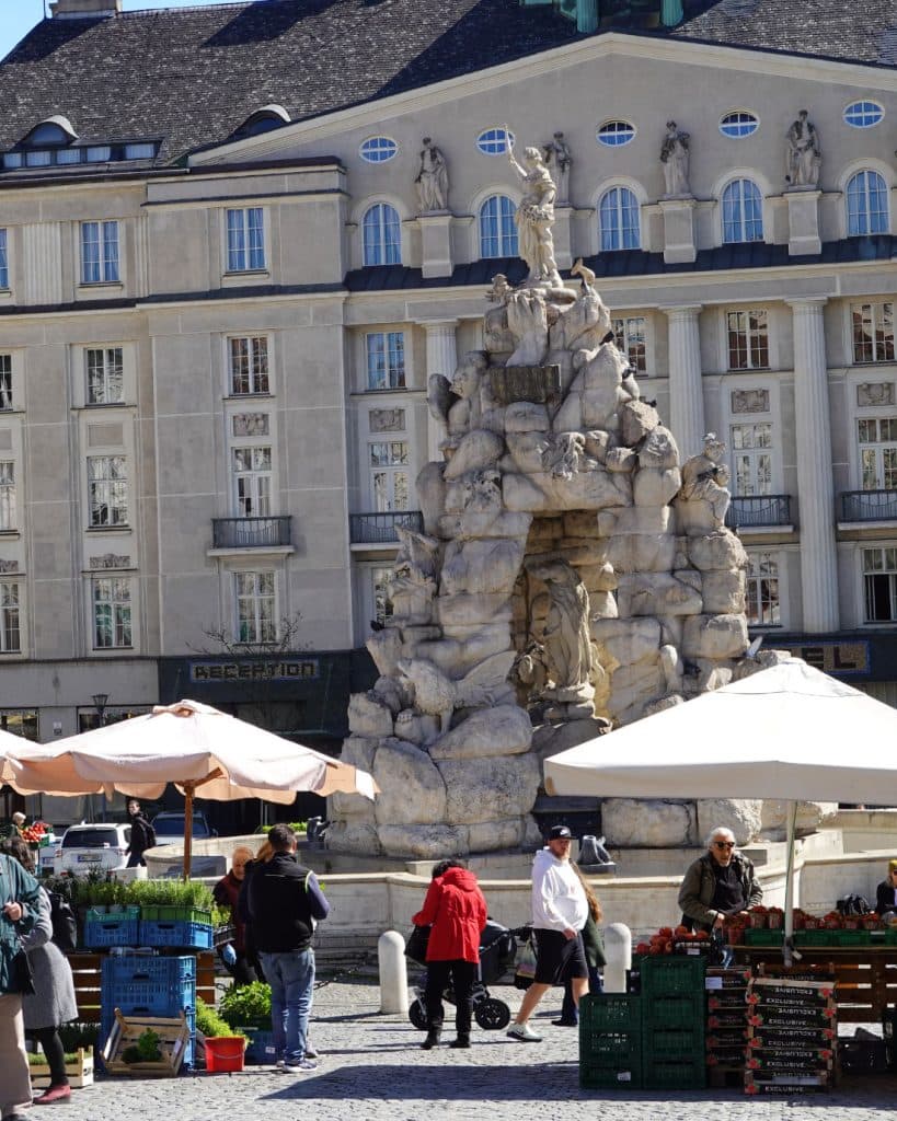 Een drukke openluchtmarkt in Brno, Tsjechië, met winkelende mensen bij een grote stenen fontein met standbeelden. Witte paraplu's geven schaduw aan marktkraampjes met producten, voor een groots gebouw versierd met standbeelden en zuilen - een genot voor reizigers.