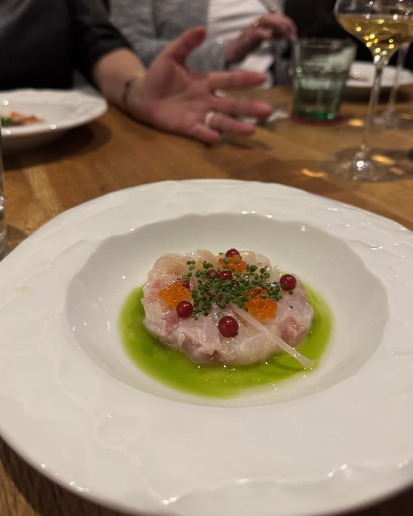 Een wit bord bevat een delicaat gerecht van fijngehakte rauwe vis met daarop rode en oranje kuit, groene kruiden en een levendige groene saus - een verleidelijk voorproefje van de Tsjechische keuken, perfect voor reisherinneringen in Brno. Op de achtergrond wachten nog meer borden.
