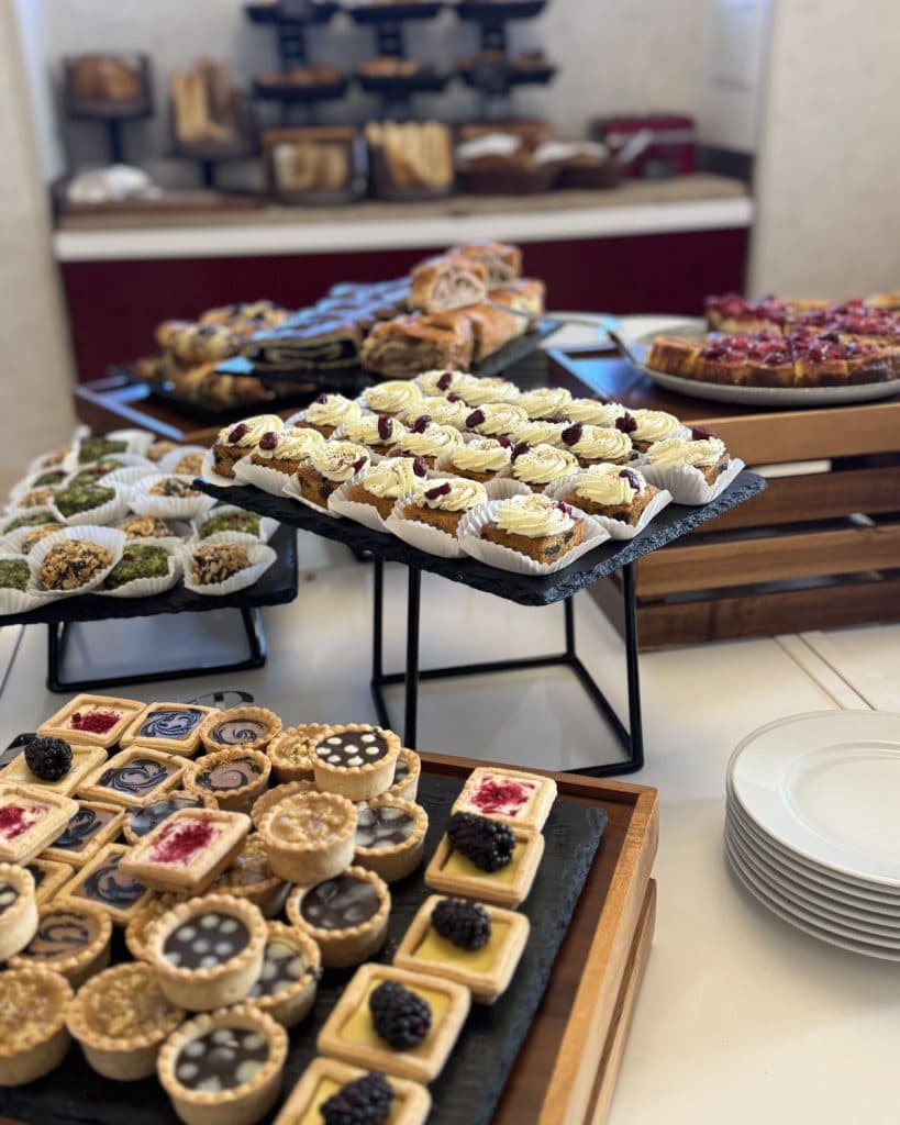 Een assortiment desserts op leistenen dienbladen, waaronder fruittaartjes, minitaartjes, pistache lekkernijen en gebak met glazuur, staat uitgestald op een buffettafel met stapelstandaards - perfect voor reizigers die Brno in Tsjechië verkennen.