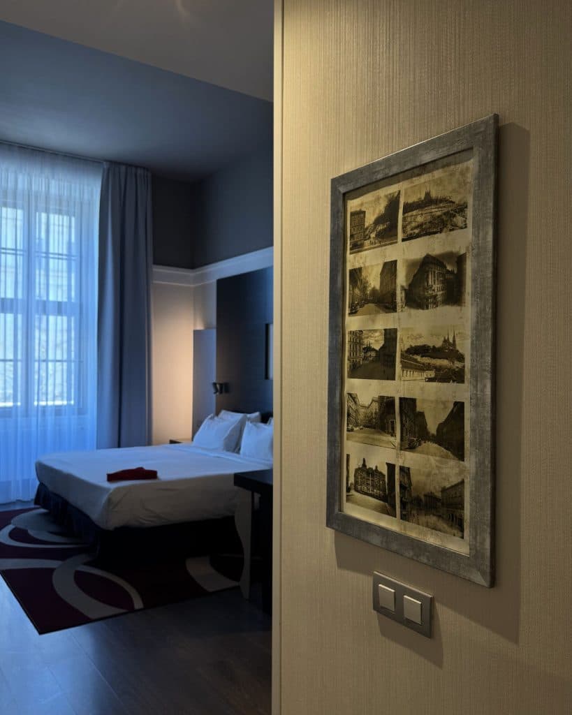 Een knusse hotelkamer in Brno, Tsjechië heeft een groot bed met wit beddengoed, sepia-getinte vintage foto's en zacht licht dat door de gordijnen naar binnen valt - een rustig toevluchtsoord voor je reisavonturen.