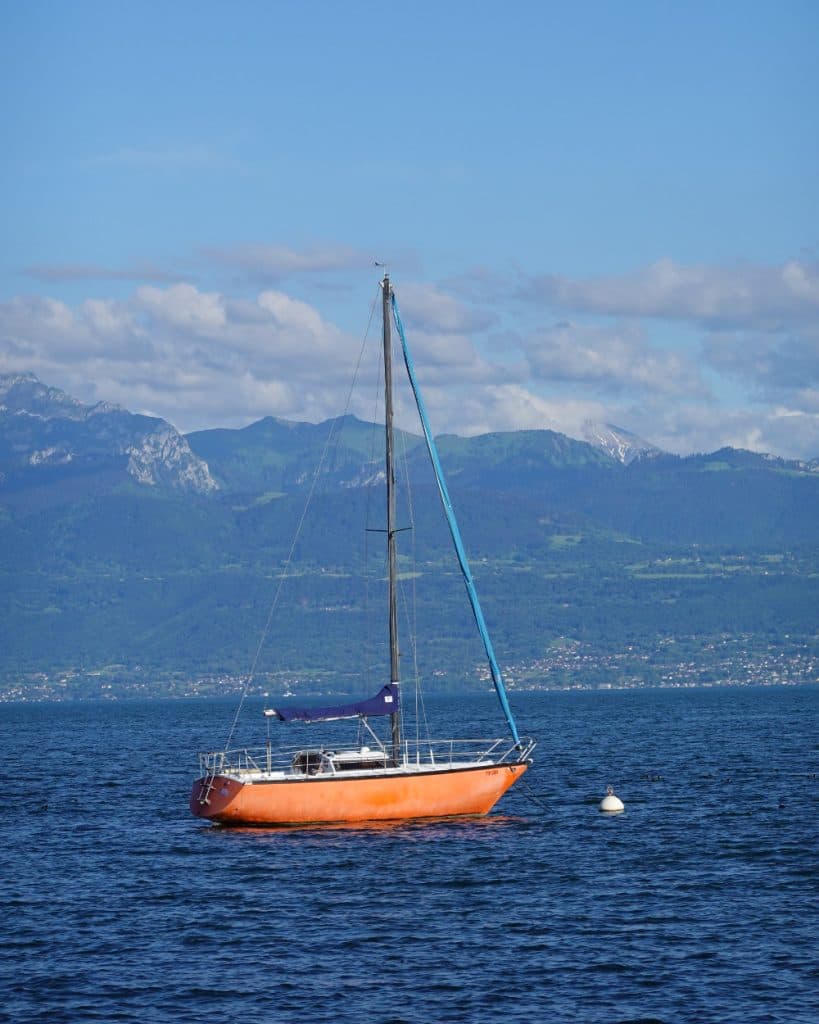 Een klein oranje zeilbootje met een blauwe mast drijft op het kalme blauwe water van het Meer van Genève bij Morges, in het kanton Vaud, vlakbij een witte boei. Op de achtergrond rijzen groene heuvels en hoge bergen op onder een blauwe hemel met hier en daar wat wolken.