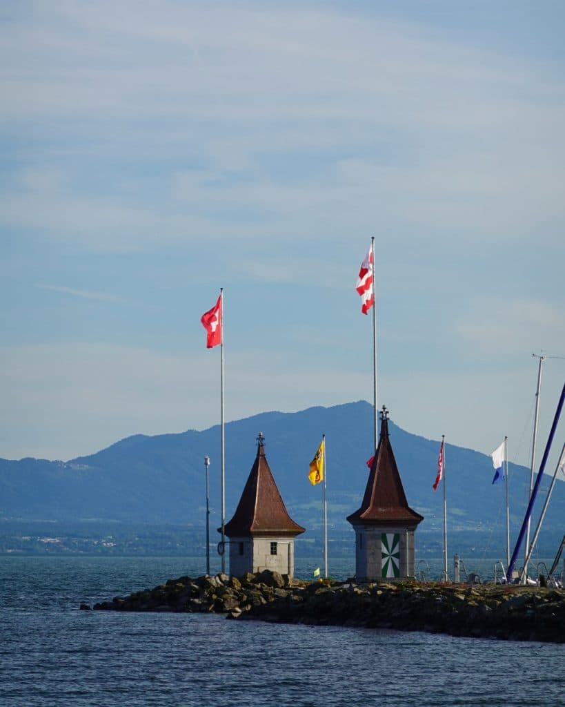 Op een rotsachtige pier aan het Meer van Genève in Morges, in het kanton Vaud, staan twee kleine stenen torentjes met puntdaken, elk met een Zwitserse vlag bovenop. Tussen beide torentjes hangt een gele vlag. Bergen en een blauwe lucht vormen het decor, met in de buurt enkele masten van zeilboten.