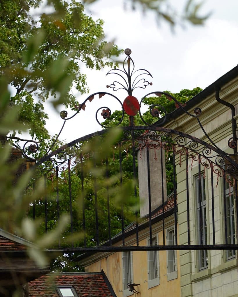 Een sierlijke smeedijzeren poort met een rood wapen bovenaan in het midden, gedeeltelijk aan het zicht onttrokken door bladrijke takken, in Morges, Vaud. Achter de poort staat een lichtgekleurd historisch gebouw met gele en witte muren, groene bomen en een bewolkte hemel.