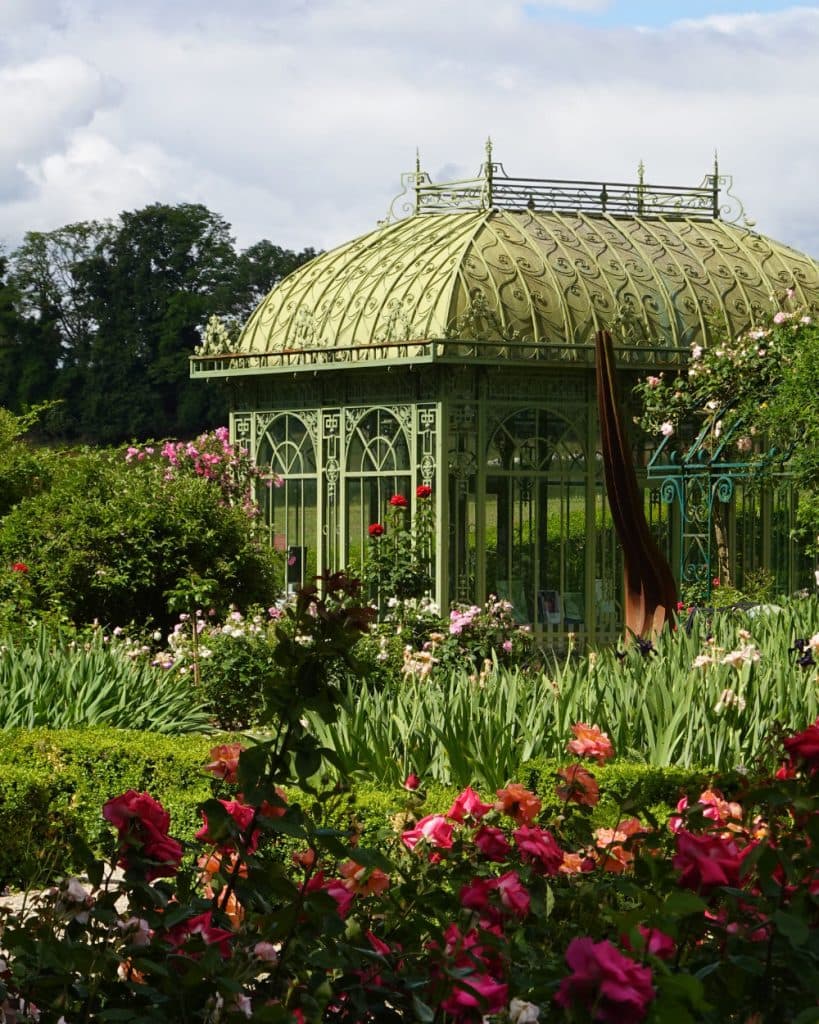 In een weelderige tuin in Morges, in het kanton Vaud, staat een sierlijk, lichtgroen prieel van smeedijzer, omringd door bloeiende rode, roze en witte bloemen. Het prieel wordt omringd door hoge groene planten en heggen, met op de achtergrond bomen en een gedeeltelijk bewolkte hemel.