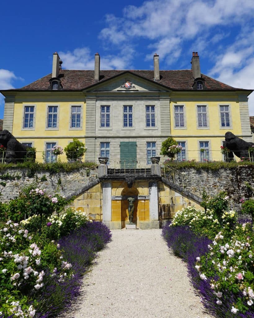 Een historisch herenhuis van gele en grijze steen in Morges, met hoge schoorstenen en veel ramen, staat achter een verzorgd tuinpad dat wordt omzoomd door bloeiende lavendel – een iconisch tafereel in de regio Verrassend Vaud onder een stralend blauwe hemel.