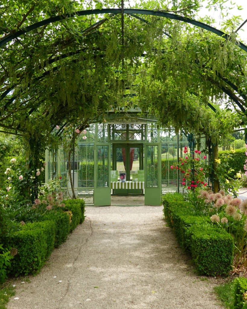 Een weelderige groene tuin in het kanton Vaud met een grindpad leidt via een met wijnranken begroeid metalen prieel naar een glazen kas. Langs het pad staan keurig gesnoeide heggen en bloeiende bloemen, en in de kas, vlakbij Morges, staat een groen-wit gestreepte bank.