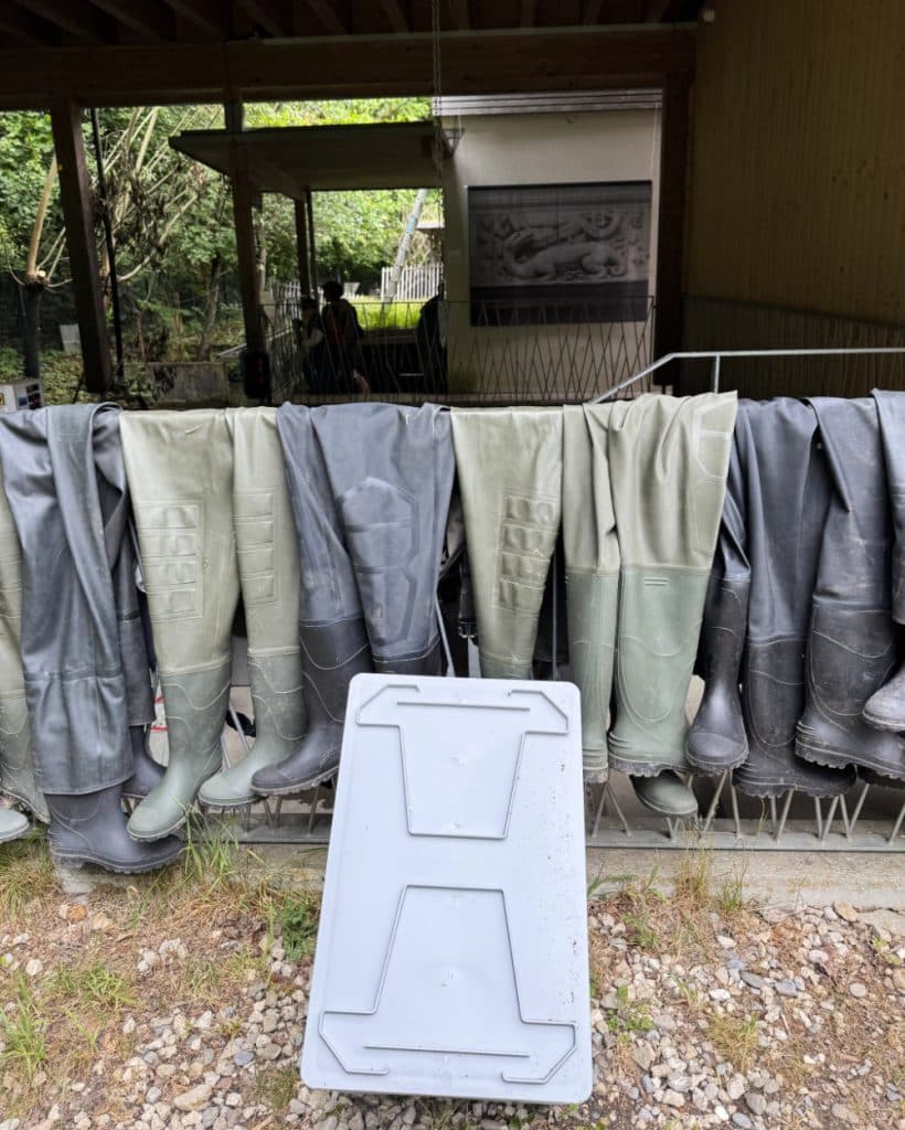 Buiten in Morges, in het kanton Vaud, hangen verschillende paar hoge rubberen waadbroeken in grijze en groene tinten ondersteboven aan een metalen rek. Daarachter bevindt zich een overdekte ruimte met een ingelijste zwart-witfoto, en op de grindgrond leunt een wit plastic deksel van een afvalbak.