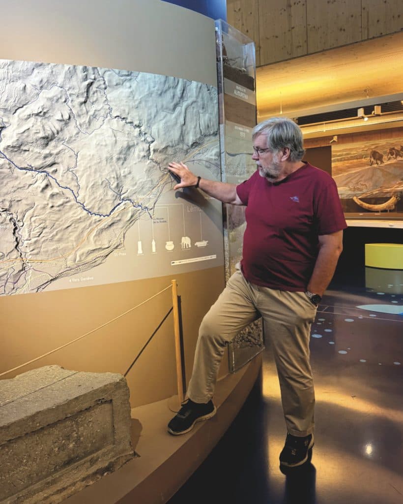 Een oudere man met grijs haar en een baard, die een bril draagt, een kastanjebruin overhemd en een beige broek, staat binnen in een museum in Morges, in het kanton Vaud, en wijst naar een grote reliëfkaart met reliëfstructuur, terwijl op de achtergrond andere tentoongestelde stukken te zien zijn.