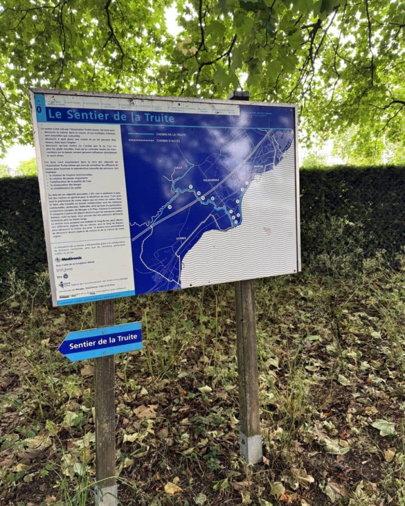 Op een grote buitenkaart met de titel „Le Sentier de la Truite“ in de buurt van Morges in het kanton Vaud staat wandelinformatie in het Frans vermeld. De kaart is bevestigd aan houten palen te midden van gras en loofbomen; een blauw pijlbordje eronder wijst naar links en draagt het opschrift „Sentier de la Truite“.