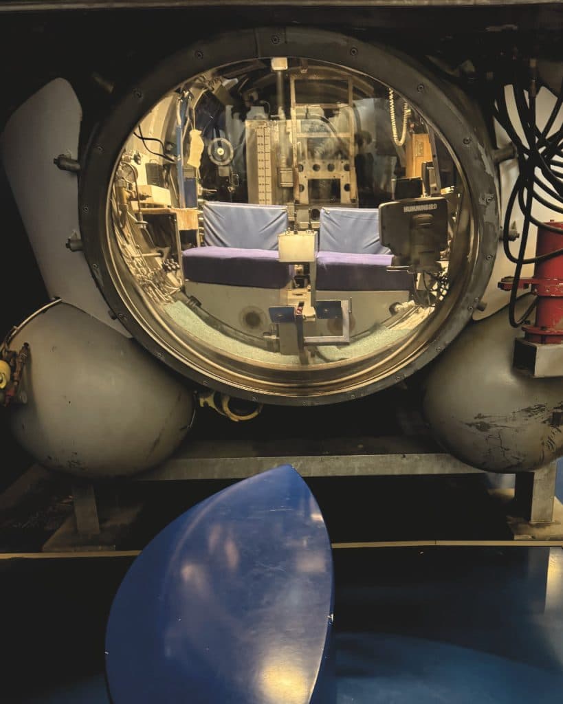 Een grote, ronde glazen kooi met een metalen frame biedt uitzicht op een ruimte met twee stoelen met blauwe kussens en bedieningspanelen, die doet denken aan een wetenschappelijk of onderwateronderzoeksapparaat — mogelijk gebruikt voor verkenningen in de meren bij Morges in Vaud.
