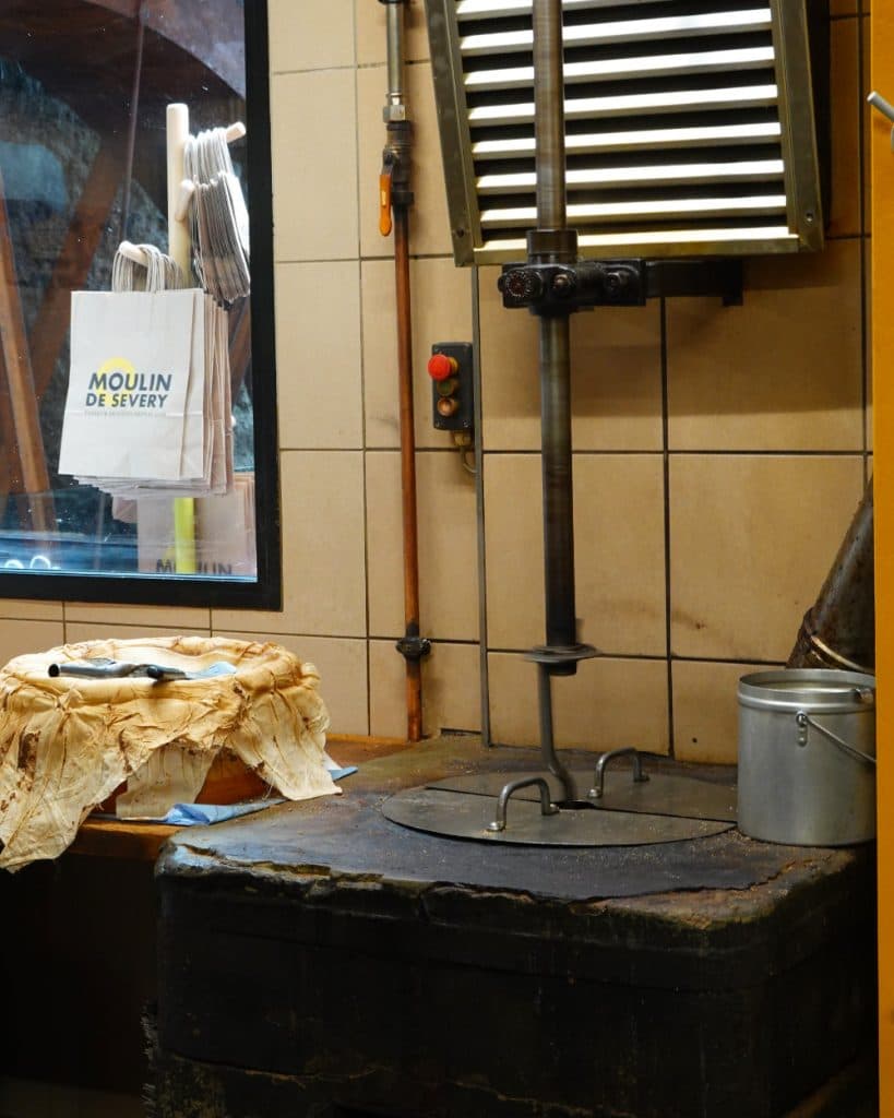 In het interieur van een oude molen in Vaud staat een verticale metalen pers op een versleten zwarte stenen sokkel, waarbij een deel van de installatie bedekt is met een beige doek. Er staat een zilverkleurige metalen emmer vlakbij. Op de achtergrond is door een raam een witte zak met het opschrift „Moulin de Sévery“ en keukengerei te zien.